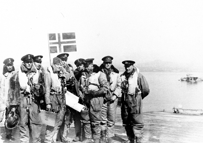 DUBLETT Norske styrker i utlandet under krigen - Det norske flyvåpen i Storbritannia. Flybåtene / sjøflyene / Catalina-flyene "som går på Norge". Mannskapet som gjorde den første turen til Norge med "Vingtor" i mai 1942. I bakgrunnen sees de norske flagget som er laget av blikk, og som er tatt med for anledningen. Like under flagget står stasjonens sjef, kaptein Lambrechts, nr. to f.h. er Nordahl Grieg, som var med på denne turen. I bakgrunnen er Catalina-flyet "Vingtor" fortøyd. WW2 - The Norwegian Air Force in Great Britain.